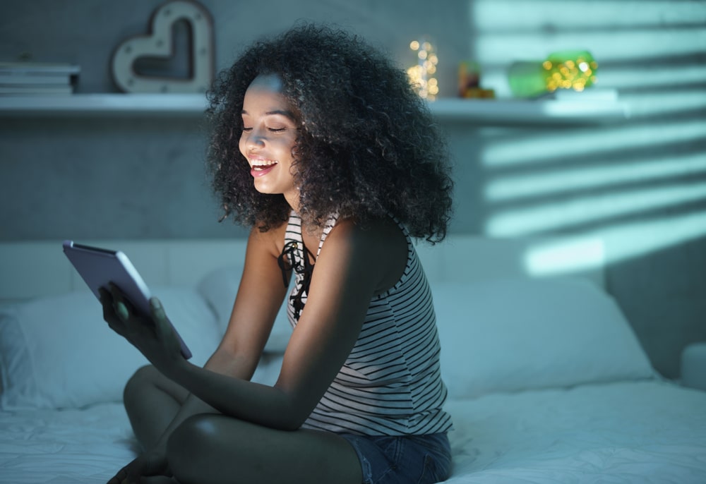 Jovem mulher sentada em uma cama olha com alegria o tablet