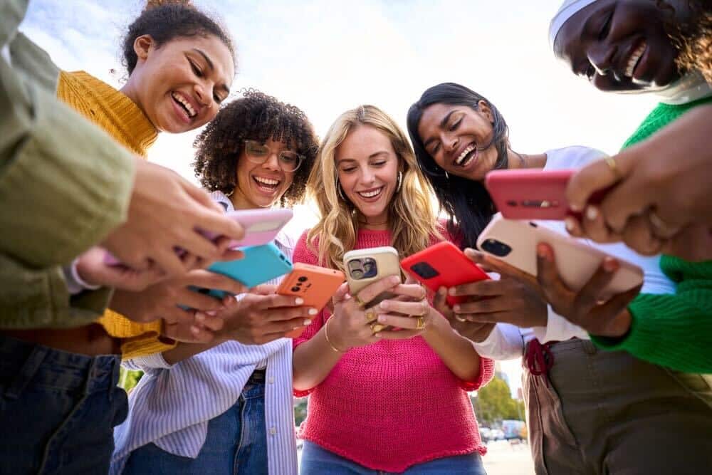 Pessoas diversas em um circulo seguram celulares com capas coloridas para mexer em Redes Sociais